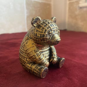 May include: A decorative bear figurine in a sitting position. The bear is gold and brown with a woven pattern design. The figurine has small ears, a snout, and detailed paws. The bear is sitting on a red surface.