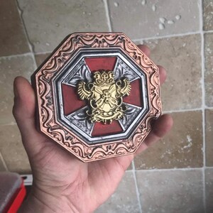 May include: A decorative octagonal shield with a red cross and a gold crest featuring a double-headed eagle. The shield has a copper-colored frame with intricate details.
