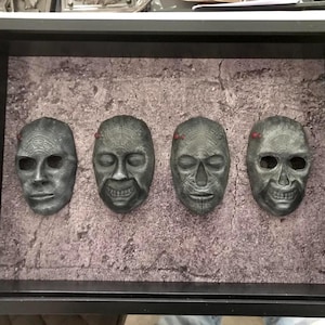 May include: A framed display of four grey, weathered masks with different facial expressions. The masks, with varying features, are set against a textured background. Small red details are visible on some masks.