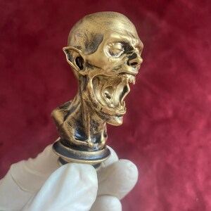 Può includere: Una scultura dorata e dettagliata della testa di una creatura con bocca aperta, denti affilati e orecchie a punta. La scultura ha una base scura ed è tenuta da una mano guantata su sfondo rosso.