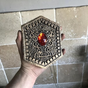 May include: A bronze-colored, hexagonal pendant with intricate embossed designs and a large, faceted, red gemstone at its center. The pendant has a chain attached and is held in a hand.