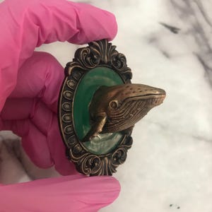May include: A bronze whale sculpture, with detailed features, emerging from a green oval, set within a dark, ornate frame. The frame has a vintage design. The sculpture is held by a person wearing a pink glove.