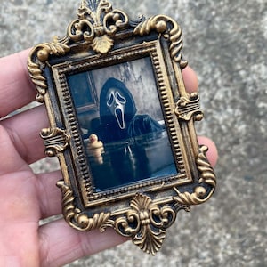 May include: Ornate gold-colored picture frame with a black and white image of the Ghostface character from the Scream horror film franchise. The frame has intricate scrollwork and a small rubber duck in the image.