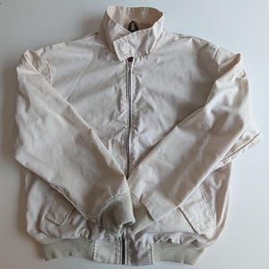 May include: A light beige bomber jacket with a zip-up front, a collared neckline, and ribbed cuffs and hem. The jacket has two front pockets and a simple, classic design. The jacket is a neutral color, suitable for various outfits.