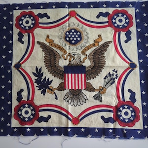 May include: A blue and white fabric square with a red, white, and blue American eagle design. The eagle is holding a bundle of arrows in one talon and an olive branch in the other. The eagle is surrounded by a wreath of stars and the words "E PLURIBUS UNUM".