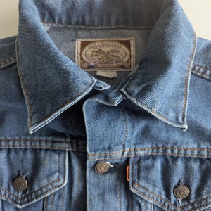 May include: Close-up of a blue denim jacket with a collar and two chest pockets. The jacket has metal buttons and orange stitching. A label inside the collar reads "Levi Strauss & Co. San Francisco, Cal."