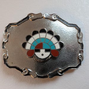 Könnte beinhalten: Eine silberfarbene Gürtelschnalle mit einem zentralen Design, das ein stilisiertes Gesicht mit Kopfschmuck zeigt. Der Kopfschmuck besteht aus schwarzen, weißen, türkisfarbenen und roten Elementen. Die Schnalle hat einen dekorativen, gewellten Rand.