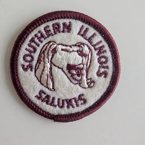 Op de afbeelding: Ronde patch met een witte kern en een bordeauxrode rand. De teksten "SOUTHERN ILLINOIS" en "SALUKIS" zijn geborduurd in bordeauxrood. Een gestileerde afbeelding van een hond staat in het midden, eveneens in bordeauxrood en wit.