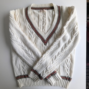 May include: Cream-colored cable knit sweater with a V-neck and long sleeves. The neckline, cuffs, and hem feature a striped design in red, gray, and brown. The sweater is folded neatly on a white surface.