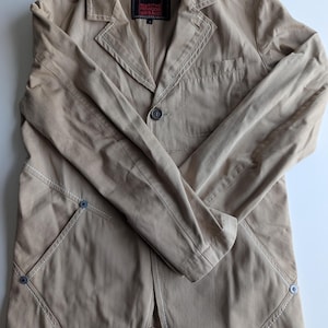 Puede incluir: Blazer color beige con costuras blancas y cierre de un botón. Cuenta con dos bolsillos delanteros y una etiqueta que dice "MARITHE FRANCOIS GIRBAUD". La chaqueta tiene un diseño clásico y entallado.