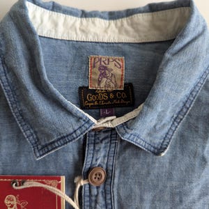 May include: A light blue denim shirt with a white collar and a brown button. The shirt has a label that reads "PRPS Goods & Co.  The Elements Made Stronger".