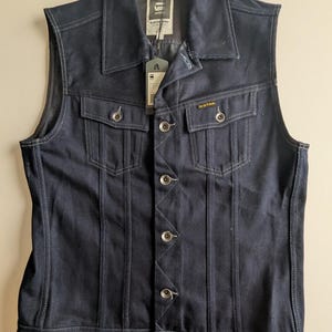 May include: A dark blue denim vest with a collared neckline and button-up closure. The vest features two chest pockets and a G-Star Raw tag. White stitching accents the seams and pockets. The vest is sleeveless.