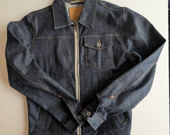 May include: A dark blue denim jacket with a zip-up front, a small chest pocket, and buttoned cuffs. The jacket has a classic collar and a small leather tag on the back of the neck.