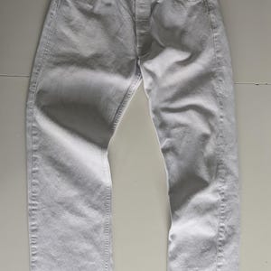 Pode incluir: Um par de calças de ganga brancas com braguilha de botões e design clássico de cinco bolsos. As calças de ganga estão estendidas, mostrando os detalhes das costuras e a forma geral. A cor clara sugere um estilo casual.