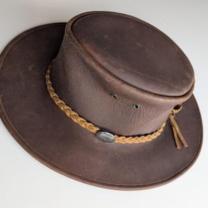 May include: A brown leather hat with a wide brim and a braided band. The hat has a distressed finish and a small metal emblem on the band. The hat has two small air vents on the side.