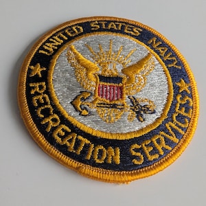 Op de afbeelding: Ronde patch met de woorden "United States Navy Recreation Services" geborduurd in goud en marineblauw. De patch heeft een centraal embleem met een gouden adelaar, een rood-wit schild en gekruiste ankers.