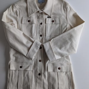 Peut inclure: Veste en jean crème avec un col, une fermeture boutonnée et quatre poches avant. La veste présente des boutons marron et des détails de couture. Le logo Wrangler est visible. Manches longues avec revers.