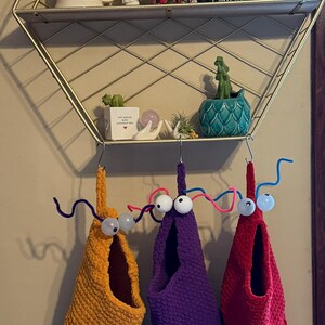 May include: Three whimsical, handmade fabric wall hangings in yellow, purple, and red, each with a fringe bottom and googly eyes. The hangings are suspended from a metal shelf with decorative items.