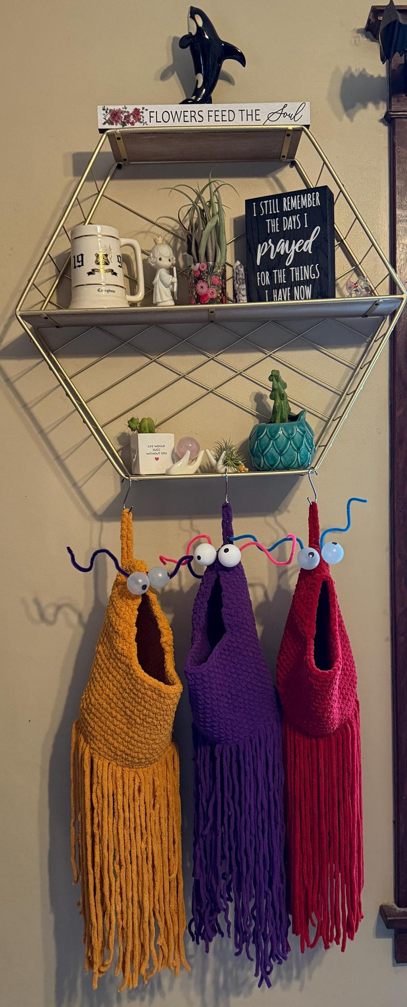 May include: Three whimsical, handmade fabric wall hangings in yellow, purple, and red, each with a fringe bottom and googly eyes. The hangings are suspended from a metal shelf with decorative items.