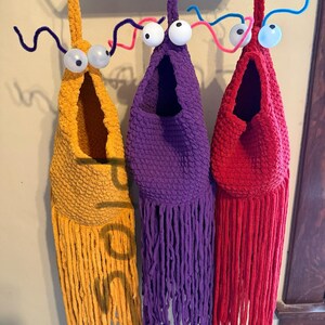 May include: Three handmade, whimsical fabric creatures in yellow, purple, and red, each with large googly eyes and pipe cleaner antennae. The creatures have open pouches and fringed bottoms, hanging from a metal rack. The word "sold" is visible on the yellow creature.