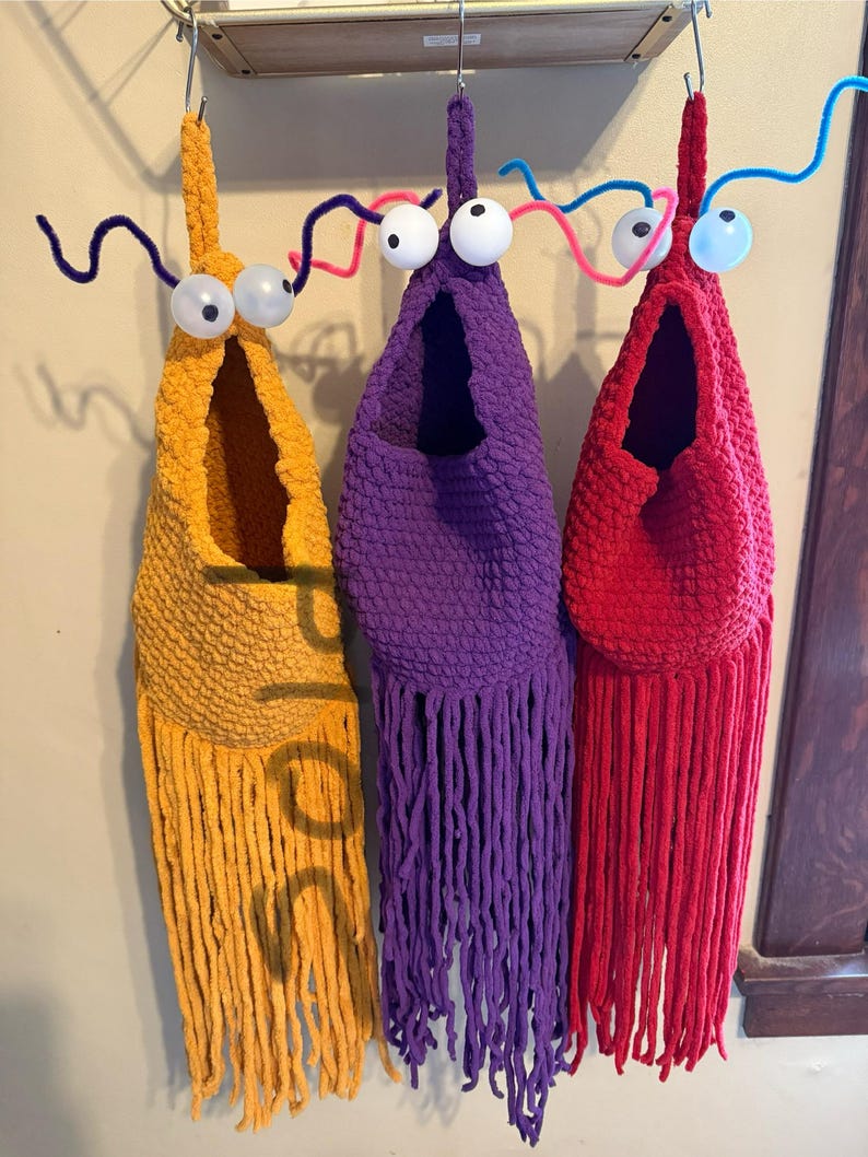 May include: Three handmade, whimsical fabric creatures in yellow, purple, and red, each with large googly eyes and pipe cleaner antennae. The creatures have open pouches and fringed bottoms, hanging from a metal rack. The word "sold" is visible on the yellow creature.