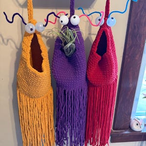 May include: Three handmade, whimsical plant holders in yellow, purple, and red, each with large googly eyes and pipe cleaner antennae. The holders are designed to hang and feature a fringe detail at the bottom. One holder contains a small air plant.