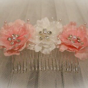 May include: A white hair comb with three pink fabric flowers and silver rhinestones. The flowers are arranged in a row on the comb.