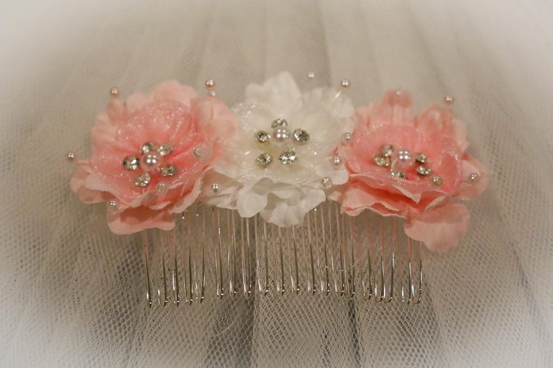 May include: A white hair comb with three pink fabric flowers and silver rhinestones. The flowers are arranged in a row on the comb.