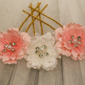 May include: Three fabric flowers with pearl and rhinestone embellishments, attached to gold bobby pins. The flowers are pink and white.