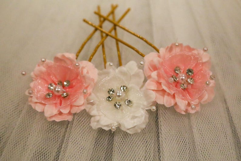 May include: Three fabric flowers with pearl and rhinestone embellishments, attached to gold bobby pins. The flowers are pink and white.