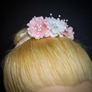 May include: A pink and white flower hair clip with rhinestones. The clip is on a blonde bun.