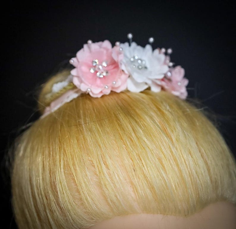 May include: A pink and white flower hair clip with rhinestones. The clip is on a blonde bun.