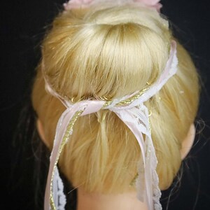 May include: A blonde hair bun with a pink and white flower hair accessory and a pink ribbon with gold trim.
