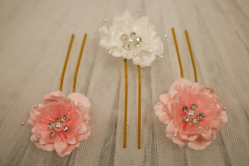 May include: Three gold hair pins with pink and white fabric flowers. Each flower has a pearl and rhinestone embellishment in the center.