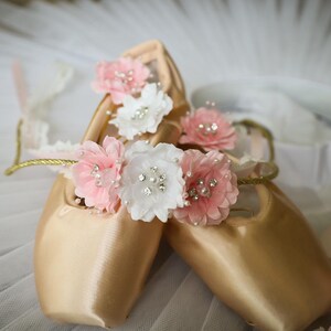 May include: A pair of gold satin ballet shoes decorated with pink and white fabric flowers. The flowers have pearl and rhinestone accents.