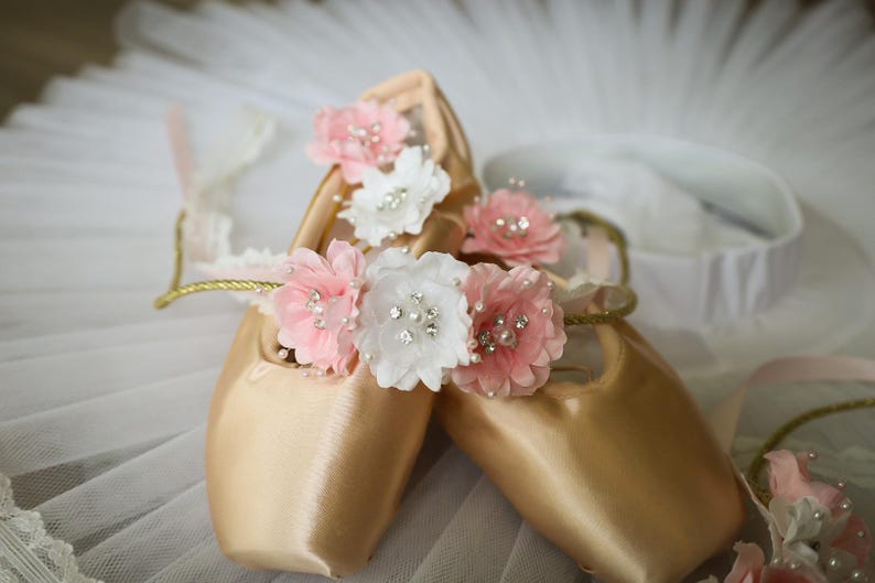 May include: A pair of gold satin ballet shoes decorated with pink and white fabric flowers. The flowers have pearl and rhinestone accents.