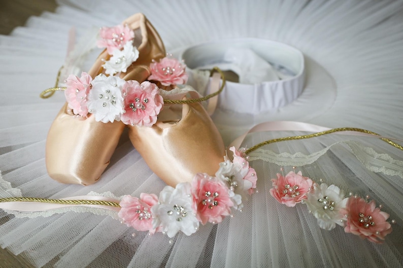 May include: Two pairs of pink ballet shoes with white and pink flower embellishments. The shoes are on a white tulle fabric.