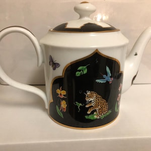 May include: White porcelain teapot with a black and gold design featuring a leopard, butterflies, and flowers. The teapot has a gold trim and a black handle.
