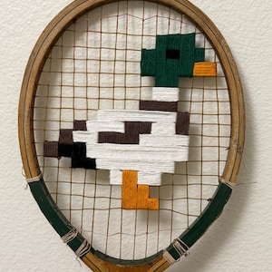 Puede incluir: Una raqueta de tenis de madera vintage con un diseño de pato pixelado tejido en las cuerdas. El pato presenta colores blanco, marrón, negro, verde y naranja. La raqueta tiene un marco marrón con un mango verde.
