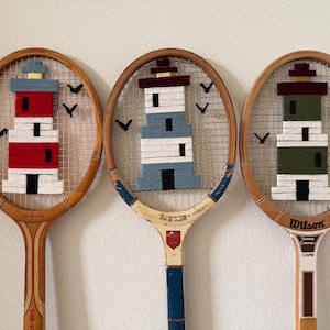 Könnte beinhalten: Drei alte Tennisschläger aus Holz mit umwickelten Leuchtturm-Motiven aus Garn. Der linke Schläger zeigt einen rot-weißen Leuchtturm, der mittlere einen blau-weißen und der rechte einen grün-weißen Leuchtturm, jeweils ergänzt durch schwarze Vogelsilhouetten.