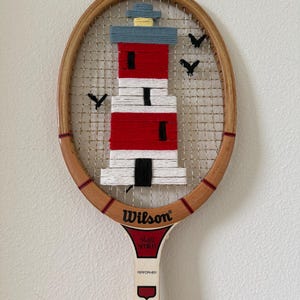 Pode incluir: Uma raquete de ténis vintage de madeira Wilson Stan Smith Performer com um design de farol feito de fios. O farol é construído com fios vermelhos, brancos, azuis e amarelos, acentuado por pássaros de fio preto na rede metálica da raquete.