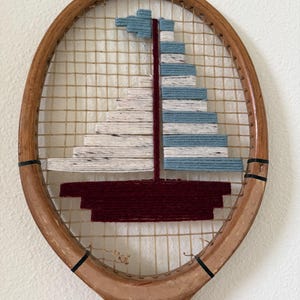 May include: A vintage wooden tennis racket featuring a decorative sailboat design woven into the strings. The boat is crafted from maroon, light blue, and speckled white yarn, creating a layered sail and hull structure within the oval frame.