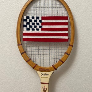 May include: Vintage Wilson Jack Kramer Autograph tennis racket with a hand-stitched American flag design. The flag features red and white stripes with a blue field of white stars. The racket has a wooden frame and a white handle.