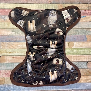 May include: A brown cloth diaper with a dark background featuring a repeating pattern of white owls, candles, and stars. The diaper has brown snaps and a brown trim.
