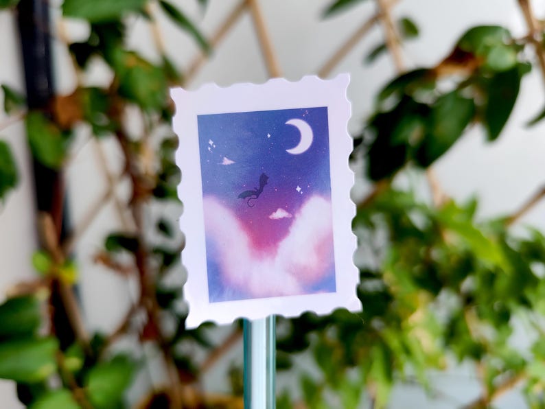May include: A decorative stamp-shaped sticker featuring a dreamy night sky scene. The design includes a crescent moon, stars, and a silhouette of a creature against a backdrop of purple and blue hues. The sticker has a white, scalloped border.