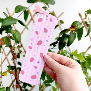 May include: A pink bookmark with a pattern of strawberries, stars, and hearts. The bookmark has vertical stripes and a pink ribbon at the top. The bookmark is held in front of a background of green leaves and a wooden trellis.