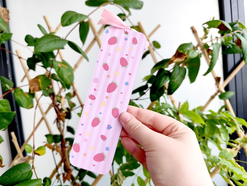 May include: A pink bookmark with a pattern of strawberries, stars, and hearts. The bookmark has vertical stripes and a pink ribbon at the top. The bookmark is held in front of a background of green leaves and a wooden trellis.