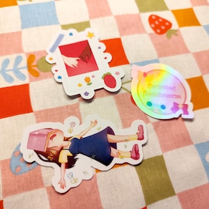 Peut inclure: Plusieurs autocollants colorés sur un tissu à carreaux. Les autocollants comprennent une image de style polaroid de deux mains qui se touchent, un autocollant holographique avec du texte et une illustration de dessin animé d'une personne allongée.