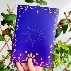 May include: A blue card with a sun design and gold decorative accents. The sun is in the center, with a starburst pattern. The card has gold floral and swirl designs around the edges.