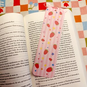 May include: A pink bookmark with a strawberry pattern, resting on an open book. The bookmark features red strawberries, yellow stars, and blue flowers. A pink ribbon is attached at the top.